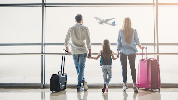 A family stands at a large window with their suitcases, watching a plane go by.